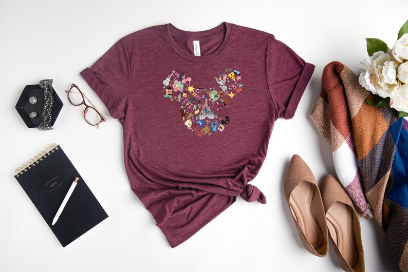 May include: A maroon t-shirt with a Mickey Mouse head design made up of various Disney characters and icons. The shirt is laid flat on a white surface.