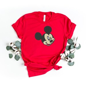 Puede incluir: Una camiseta roja con la cara sonriente de Mickey Mouse bordada en la parte delantera. El personaje cl&aacute;sico de dibujos animados aparece en la camiseta. La camiseta est&aacute; peinada con un nudo en la parte inferior y est&aacute; rodeada de vegetaci&oacute;n.