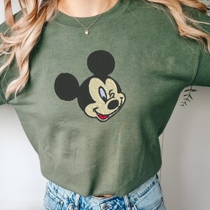 Puede incluir: Una sudadera verde oliva con un bordado de la cara de Mickey Mouse. El personaje de dibujos animados tiene la cabeza y las orejas negras, la cara beige y una sonrisa roja. La sudadera es de estilo casual.