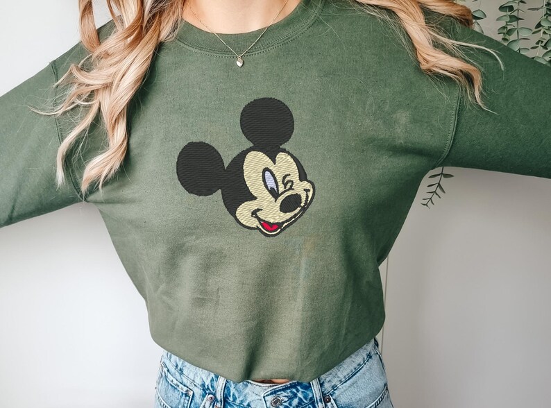 Puede incluir: Una sudadera verde oliva con un bordado de la cara de Mickey Mouse. El personaje de dibujos animados tiene la cabeza y las orejas negras, la cara beige y una sonrisa roja. La sudadera es de estilo casual.