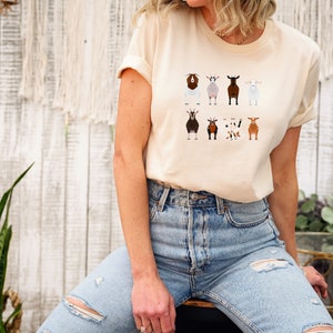 May include: A light beige t-shirt with a graphic of nine different goats in various colors and patterns. The goats are all facing forward and have a simple, cartoon-like style.