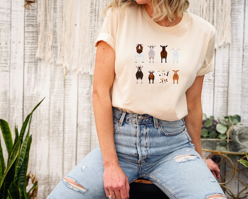 May include: A light beige t-shirt with a graphic of nine different goats in various colors and patterns. The goats are all facing forward and have a simple, cartoon-like style.