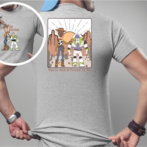 May include: A light grey t-shirt with a cartoon graphic of Woody and Buzz Lightyear in a desert scene. The text "You've Got A Friend In Me" is below the graphic. The shirt also has a smaller graphic on the front with the text "Big Brother".