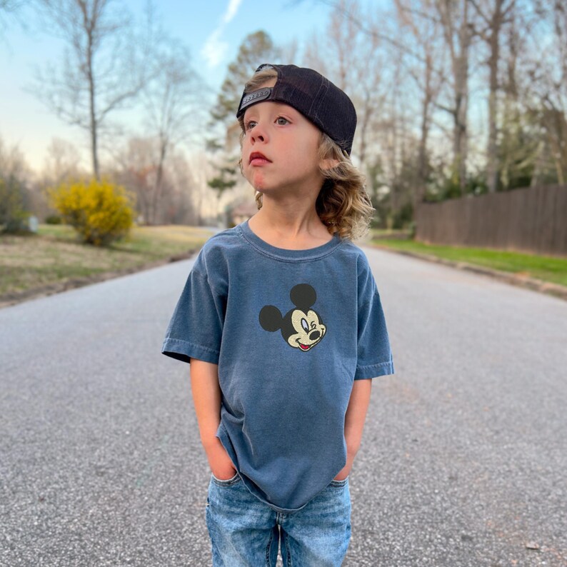 Puede incluir: Una camiseta azul con un gr&aacute;fico de Mickey Mouse, combinada con vaqueros azules y una gorra de b&eacute;isbol de malla negra. El ni&ntilde;o est&aacute; de pie en una carretera pavimentada.