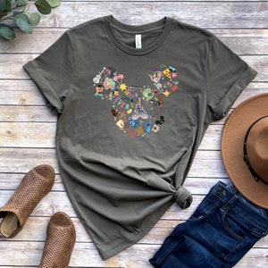 May include: A green t-shirt with a graphic of a Mickey Mouse head made up of various Disney characters and icons. The shirt is laid out on a wooden surface with a brown hat, blue jeans, and brown sandals.