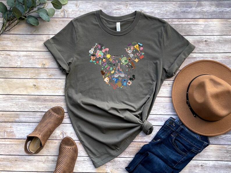 May include: A green t-shirt with a graphic of a Mickey Mouse head made up of various Disney characters and icons. The shirt is laid out on a wooden surface with a brown hat, blue jeans, and brown sandals.
