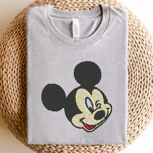 Puede incluir: Camiseta gris claro con un bordado de Mickey Mouse. El dise&ntilde;o incluye orejas negras, una cabeza negra y una cara sonriente con boca roja, hocico beige y ojos azules. La camiseta est&aacute; hecha de material suave.