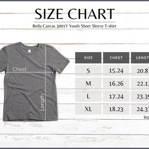 Pode incluir: Uma camiseta cinza de manga curta juvenil Bella Canvas 3001Y com uma tabela de tamanhos. A tabela mostra as medidas do peito e comprimento em polegadas para os tamanhos S, M, L e XL. A camiseta est&aacute; em um fundo de madeira branca.