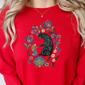 May include: A red sweatshirt featuring a black bear and a girl holding hands, surrounded by a floral design in blue, green, and yellow. The design is centered on the front of the sweatshirt.