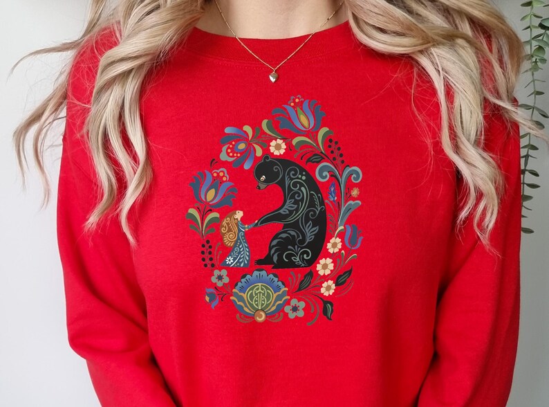 May include: A red sweatshirt featuring a black bear and a girl holding hands, surrounded by a floral design in blue, green, and yellow. The design is centered on the front of the sweatshirt.