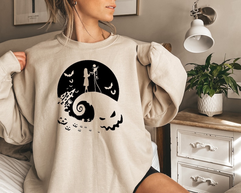 K&ouml;nnte beinhalten: Ein beiger Pullover mit einem schwarzen Grafikdruck von Jack Skellington und Sally aus Nightmare Before Christmas. Die Grafik zeigt sie auf einem H&uuml;gel stehend, im Hintergrund eine gruselige Halloween-Stadt.