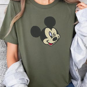 Puede incluir: Camiseta verde oliva con una imagen bordada de la cara de Mickey Mouse. El dise&ntilde;o incluye orejas negras, una cara color crema y una boca roja. La camiseta de manga corta est&aacute; hecha de un material suave.