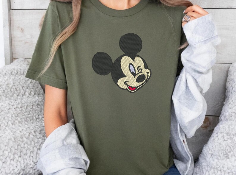Puede incluir: Camiseta verde oliva con una imagen bordada de la cara de Mickey Mouse. El dise&ntilde;o incluye orejas negras, una cara color crema y una boca roja. La camiseta de manga corta est&aacute; hecha de un material suave.