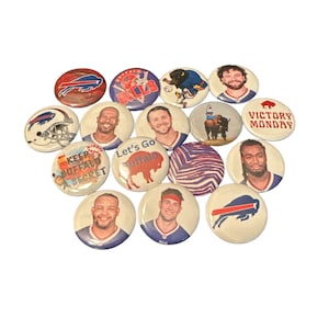 May include: A collection of 14 round buttons featuring various designs related to the Buffalo Bills football team. The buttons feature images of players, logos, and slogans like "Let's Go Buffalo" and "Victory Monday".