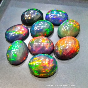 Könnte beinhalten: Eine Sammlung von neun ovalen schwarzen Opalen mit schillernden Regenbogenfarben. Die Opale sind in einem Halbkreis auf einer grauen Oberfläche angeordnet.