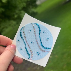 May include: A circular light blue patch decorated with curved lines of small, reflective teal and iridescent rhinestones. The design features a textured fabric base with scattered individual crystals arranged in a wave-like pattern across the surface.