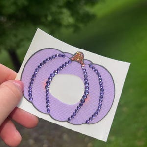 May include: A light purple pumpkin-shaped sticker or patch featuring vertical rows of small, shimmering purple rhinestones and a small, glittery orange stem at the top, all set against a white rectangular backing.