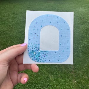 Può includere: Una toppa in tessuto azzurro a forma di lettera O, caratterizzata da una trama testurizzata e decorata con una sfumatura di piccoli strass blu e trasparenti scintillanti, densamente raggruppati nell'angolo in basso a sinistra.
