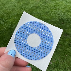May include: A circular light blue fabric patch featuring a center cutout and a dense pattern of small, reflective blue and clear rhinestones. The circular patch is attached to a square white paper backing for application and display purposes.