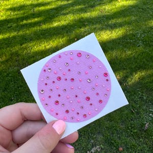 May include: A circular patch of light purple fabric decorated with an assortment of pink and clear rhinestones and beads of varying sizes scattered across the surface, attached to a square white backing card.