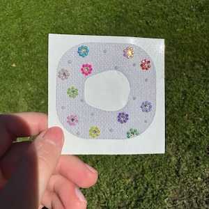 May include: A square white adhesive patch with a central circular cutout, decorated with small, colorful flower-shaped rhinestones in shades of blue, pink, green, purple, and orange, interspersed with tiny white beads on a textured fabric surface.