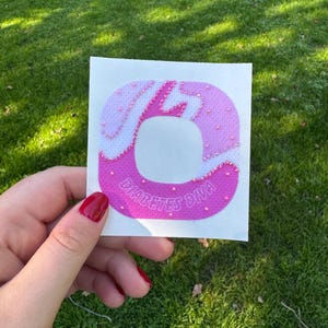 May include: A square adhesive patch featuring a circular cutout in the center. The patch has a gradient design transitioning from light pink to dark pink, decorated with small pink beads and the text DIABETES DIVA printed in white.