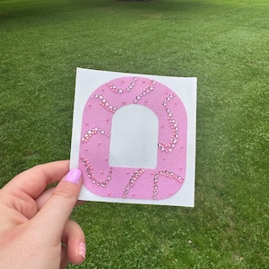 Può includere: Una toppa adesiva rosa con un ritaglio rettangolare al centro, caratterizzata da una superficie in tessuto testurizzato decorata con strass rosa sparsi e piccole perline rosa disposte in motivi curvi e vorticosi sulla superficie anteriore.