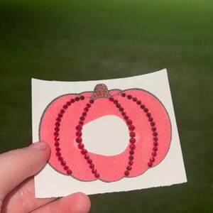 May include: A pink pumpkin-shaped iron-on patch featuring a dark outline and vertical rows of small, reflective red rhinestones. The pumpkin has a small stem at the top and is printed on a white rectangular backing material.