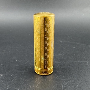 Könnte beinhalten: Ein zylindrisches Lippenstiftgehäuse aus goldfarbenem Metall mit einem vertikalen, sich wiederholenden Muster aus kleinen eingravierten Symbolen auf dem Körper. Die Basis der Hülse ist mit dem Text "TRUE-COLOR" in Großbuchstaben geprägt.