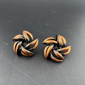 May include: A pair of circular earrings featuring a swirling, multi-layered design. Each earring is composed of curved, petal-like segments that alternate between a dark, oxidized metallic finish and a warm, burnished copper-toned surface.