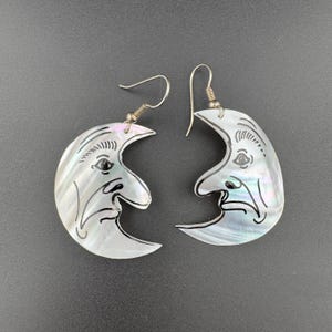 May include: A pair of iridescent mother-of-pearl earrings shaped like crescent moons. Each earring features a carved face with black etched details, including an eye, nose, and mouth, suspended from silver-toned metal ear wires for pierced ears.