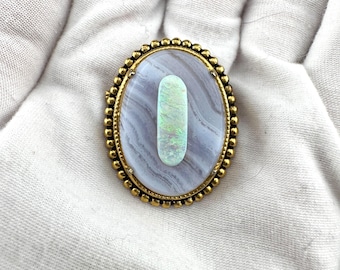 May include: An oval brooch featuring a central vertical cabochon of iridescent white opal set against a smooth, light gray banded agate stone. The piece is framed by a textured gold-toned metal border with a repeating beaded edge design.