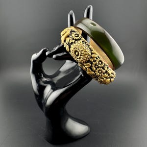 May include: Two bangles displayed on a black hand-shaped stand. One is a dark green, smooth band. The other is gold-colored, intricately carved with floral patterns. The stand is glossy and the background is dark.