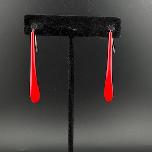 May include: A pair of long, slender, bright red drop earrings with a smooth, glossy finish. Each earring features a tapered, teardrop-like shape suspended from a thin metal hook, showcasing a vibrant solid color and a minimalist, elongated design.
