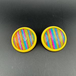 May include: A pair of round clip-on earrings featuring a bright yellow border and a central circular insert decorated with vertical stripes in vibrant shades of orange, blue, pink, and green. The surface of the center has a faceted texture.
