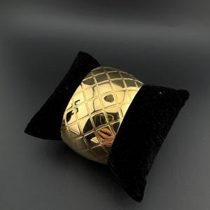 Può includere: Un bracciale a polsino largo color oro con un motivo geometrico a griglia. Il bracciale è esposto su una superficie di velluto nero, creando un contrasto che ne evidenzia la finitura lucida. Il design del bracciale è un pezzo audace.