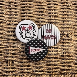Könnte beinhalten: Drei runde Ansteckbuttons. Ein weißer Button zeigt eine schwarze Bulldoggen-Illustration mit roten sternförmigen Sonnenbrillen. Ein weiterer weißer Button hat schwarze vertikale Streifen und den roten Text GO DAWGS. Ein schwarzer Button hat weiße Punkte und einen roten Wimpel mit dem Text GEORGIA.