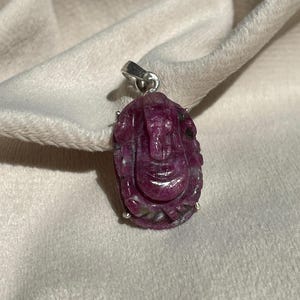 Puede incluir: Un colgante de rubí tallado con una figura de deidad. El colgante es de color púrpura rojizo intenso con detalles plateados y una fianza plateada. El colgante está sobre un fondo suave y claro.