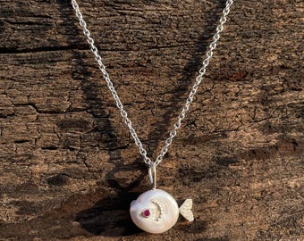 Puede incluir: Un collar de plata con un colgante de pez. El colgante es una perla blanca con un pequeño ojo de gema roja y una fila curva de pequeñas gemas blancas. El pez tiene una pequeña aleta plateada.