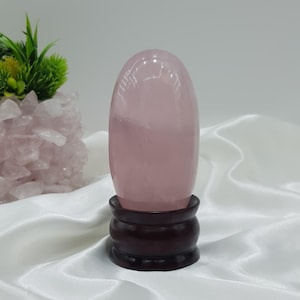 May include: A smooth, oval rose quartz crystal, in a soft pink hue, is displayed on a dark wooden stand. The crystal's surface is glossy. A small plant and a cluster of rose quartz crystals are visible in the background.