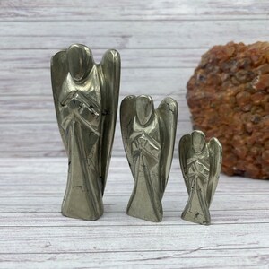 May include: Three pyrite angel figurines of varying sizes. The angels are carved from a silvery-grey stone with a metallic sheen. The largest angel is about 6 inches tall, the middle angel is about 4 inches tall, and the smallest angel is about 2 inches tall.