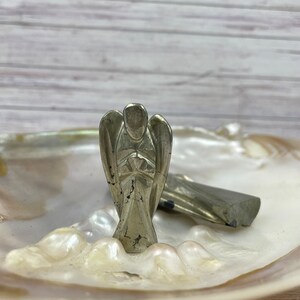 May include: A silver angel figurine carved from pyrite, a metallic mineral, is displayed on a white iridescent shell. The angel is standing with its hands clasped in front of its chest.