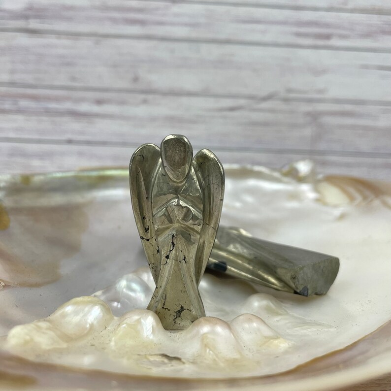 May include: A silver angel figurine carved from pyrite, a metallic mineral, is displayed on a white iridescent shell. The angel is standing with its hands clasped in front of its chest.
