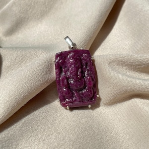 Puede incluir: Un colgante rectangular de color magenta intenso con una imagen tallada de una deidad. El colgante tiene una fianza plateada y está enmarcado con pequeños detalles plateados. La figura tallada es detallada y destaca sobre la piedra pulida.