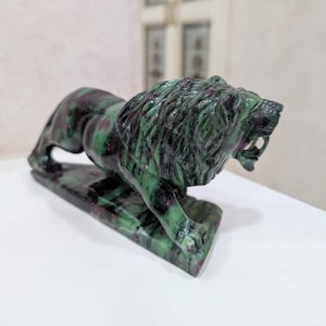May include: A carved stone lion sculpture in shades of green and red. The lion is in a crouching position with its mouth open, revealing its teeth. The sculpture is on a rectangular base.
