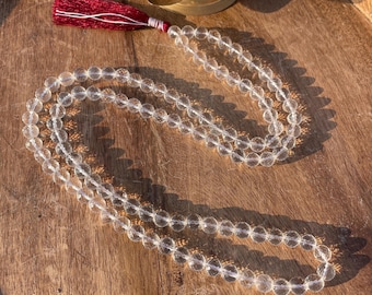 May include: A clear quartz mala with a red tassel. The mala is made of round beads and is strung on a thin cord. The mala is lying on a wooden surface.