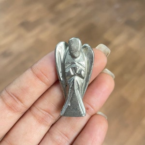 May include: A silver angel figurine carved from pyrite, a metallic mineral. The angel is standing with its arms crossed in front of its chest.