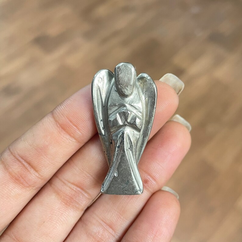 May include: A silver angel figurine carved from pyrite, a metallic mineral. The angel is standing with its arms crossed in front of its chest.