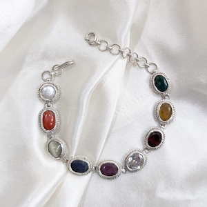 May include: A silver bracelet featuring a series of oval-shaped gemstones in various colors, including red, green, and purple. The gemstones are set in a silver frame with a twisted rope design. The bracelet is displayed on a white, silky fabric.