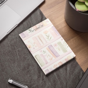 May include: A rectangular notepad with a watercolor design of bookshelves, flowers, and books in pastel colors. The word "Personalize" is written at the top. The notepad is on a desk next to a laptop and a pen.